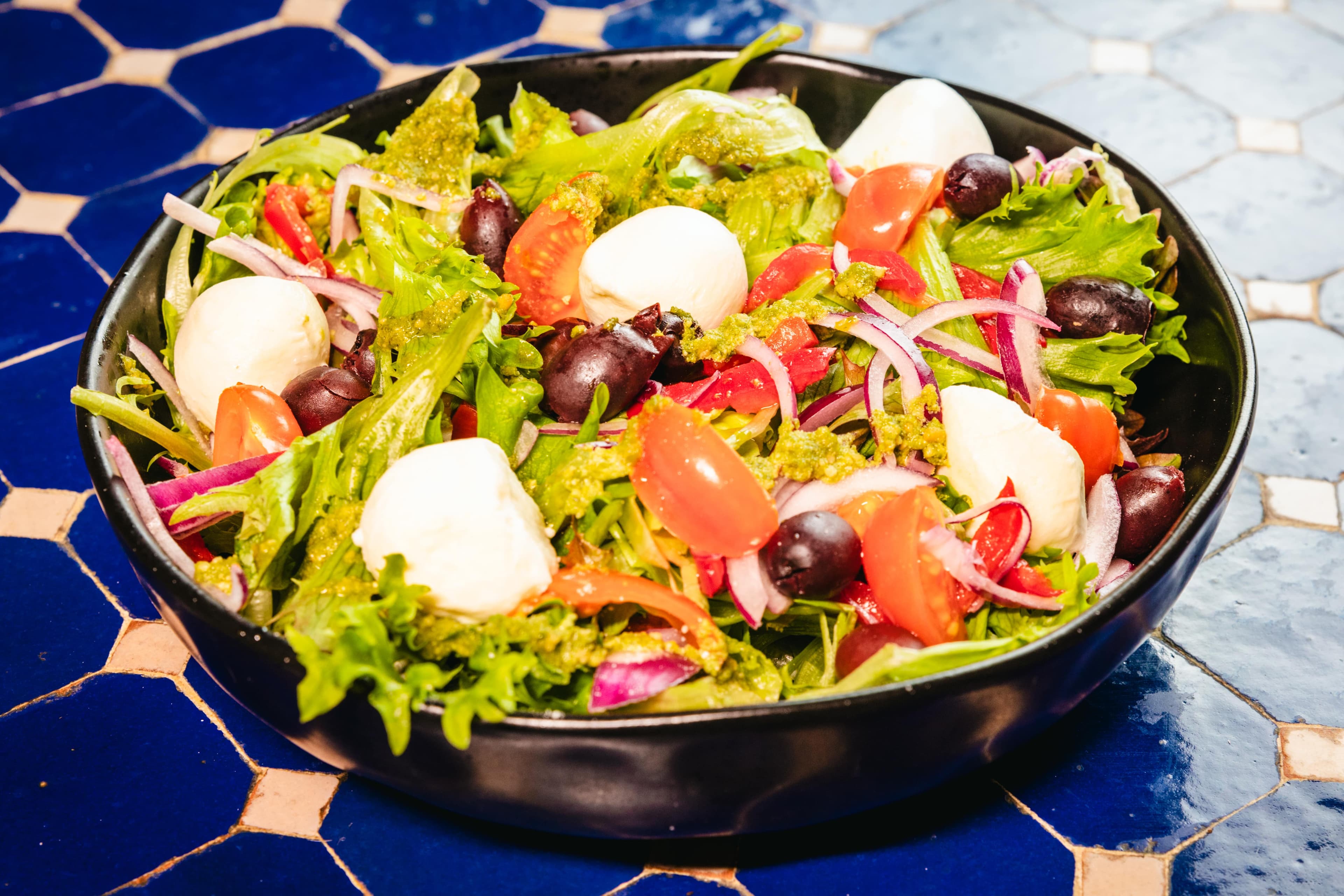Fresh salad with mozzarella, tomatoes, olives, and pesto in a black bowl on blue tiles.