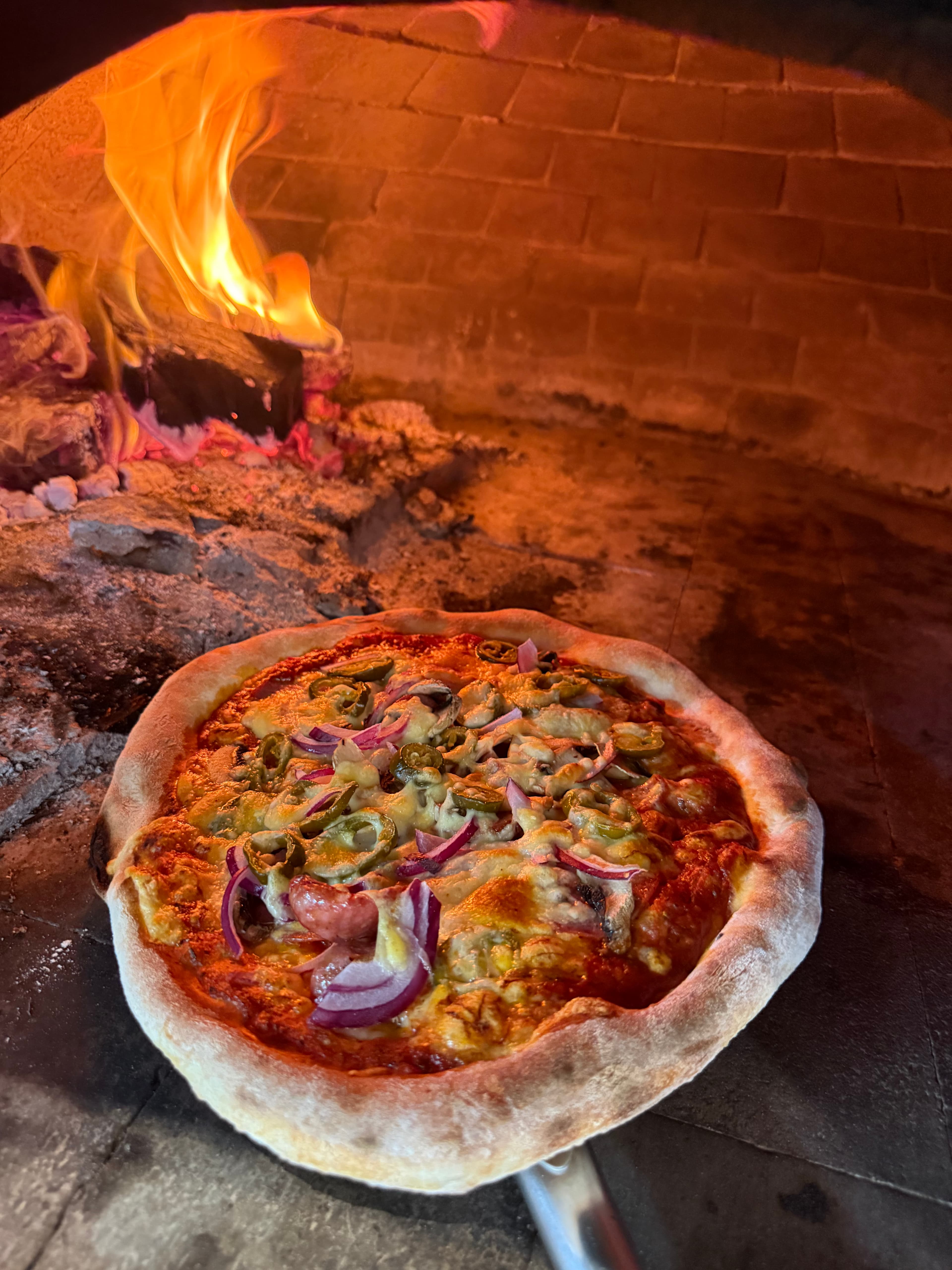 Pizza with red onions and jalapeños baking in a wood-fired brick oven with bright flames.