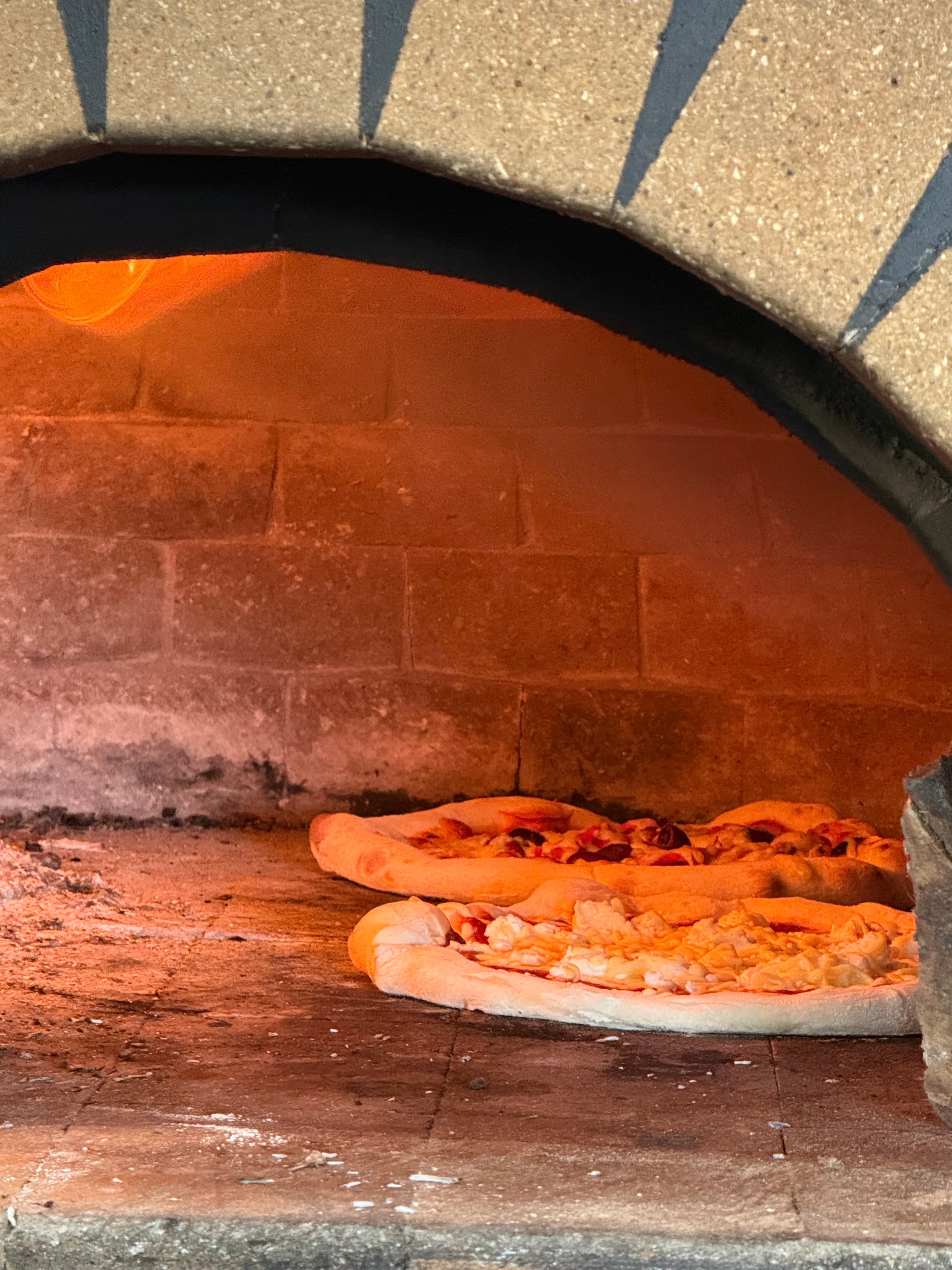 Two pizzas baking on a stone floor inside a glowing, wood-fired brick oven.